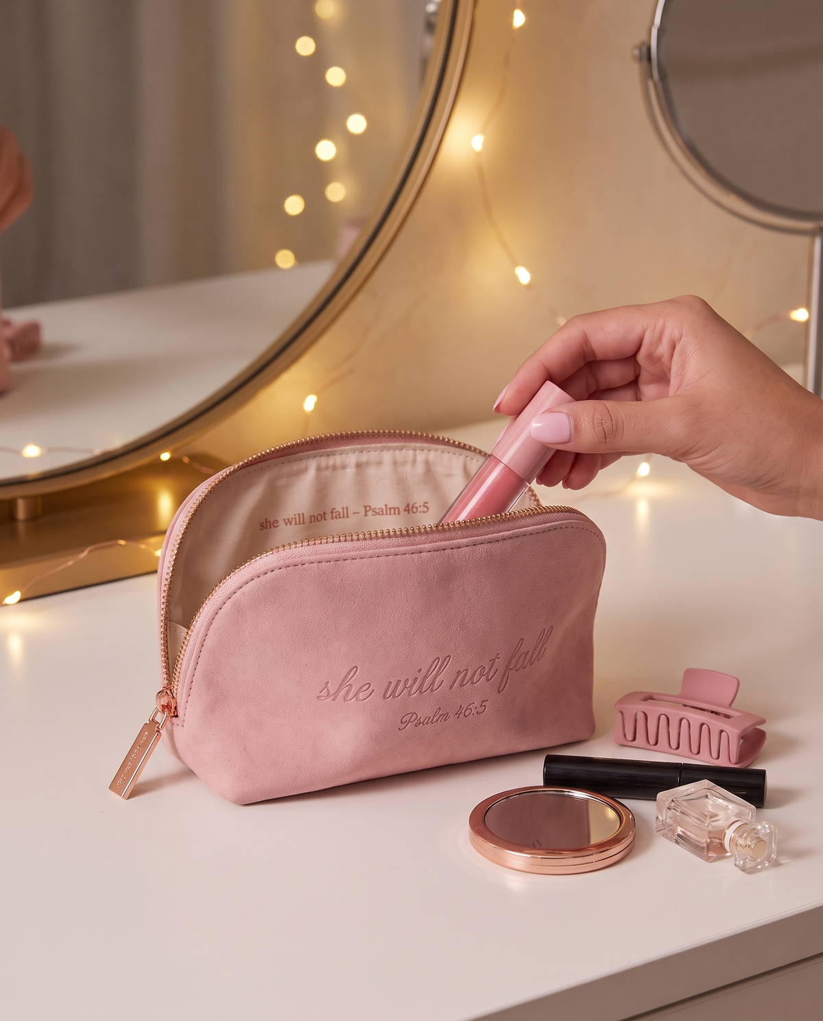 The pink Psalm 46:5 makeup pouch open on a vanity table with fairy lights